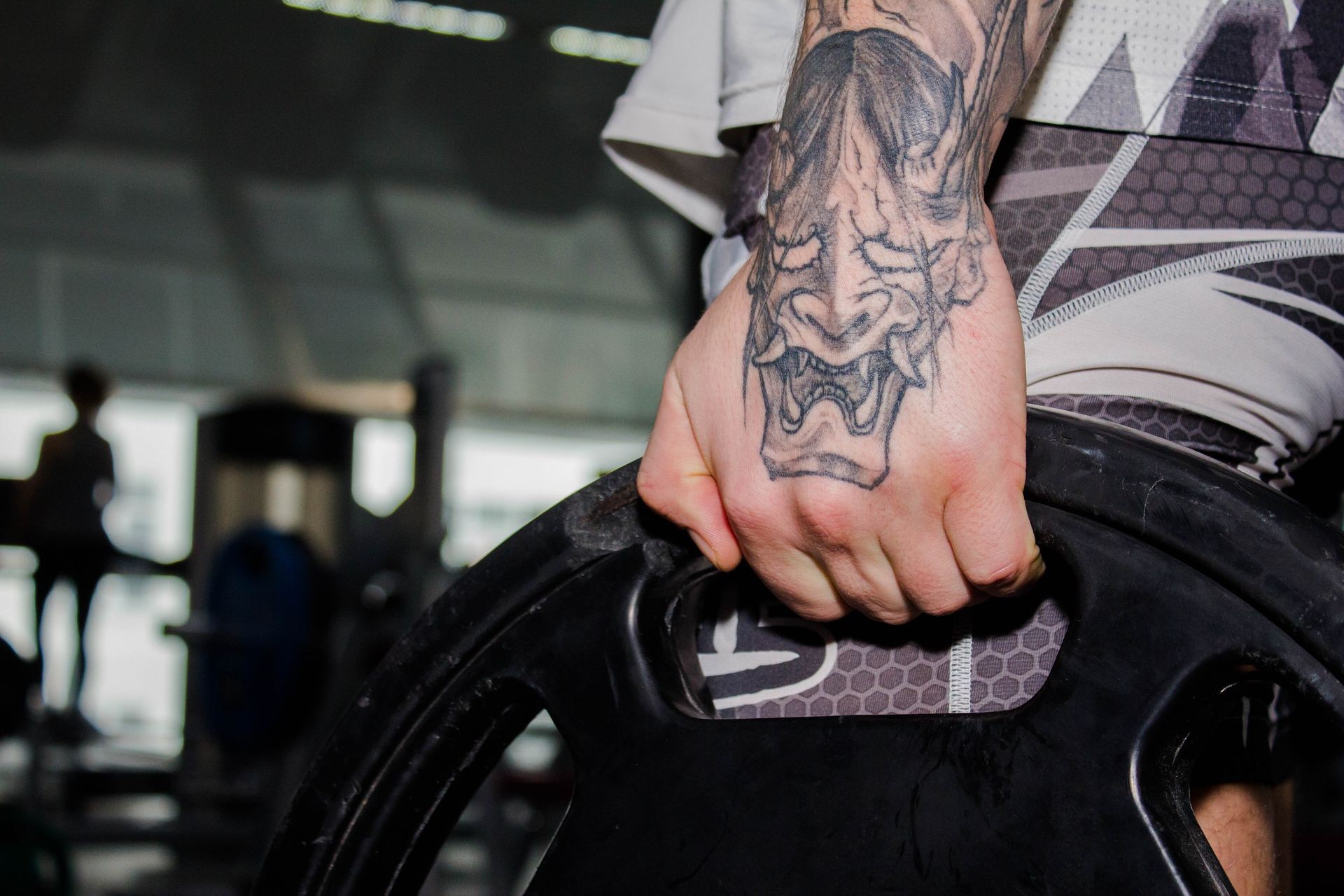 Tattoed hand of MMA young fighter holding heavy weight disc, plate for barbell in gym. MMA training program. Weightlifting in a power lifting belt, powerlifting. Lifestyle, healthy, sport. Stylish.